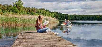VRK Premium Rente – Eine junge Frau sitzt auf einem Steg an einem See während ihr Mann mit den Töchtern in einem Boot fährt.