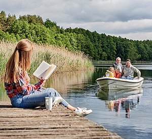 VRK Altersvorsorge – Eine junge Frau sitzt auf einem Steg an einem See während ihr Mann mit den Töchtern in einem Boot fährt.