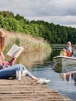 Eine junge Frau sitzt auf einem Steg an einem See während ihr Mann mit den Töchtern in einem Boot fährt.
