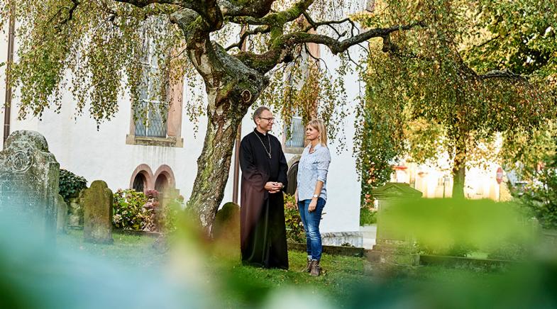 VRK Amts- und Vermögens­schaden­haftpflicht­versicherung – Ein Kirchenbeamter steht mit einer Frau vor einem Kirchengebäude.