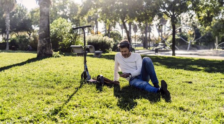 VRK E-Scooter-Versicherung – Ein Mann sitzt mit Kopfhörern neben seinem E-Scooter im Gras.