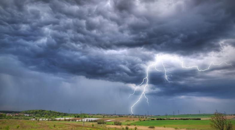  VRK Elementar­schaden­versicherung – Ein wolkenverhangener Himmel mit einem Blitzeinschlag und Regen in der Ferne.