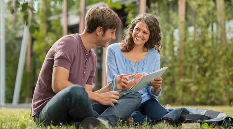 VRK Seminar-Rücktrittsversicherung – Eine junge Frau und ein Mann sitzen vor einem Gebäude im Gras und tauschen sich aus.