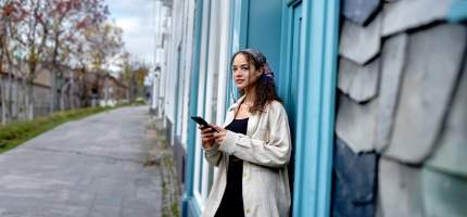 Eine junge Frau lehnt an der Haustür und blickt nachdenklich von ihrem Handy auf.