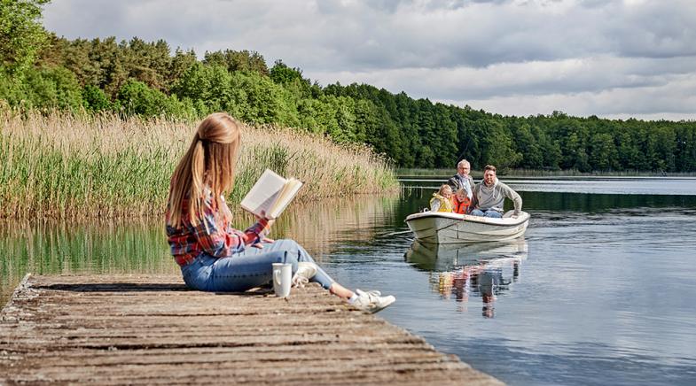VRK Premium Rente – Eine junge Frau sitzt auf einem Steg an einem See während ihr Mann mit den Töchtern in einem Boot fährt.